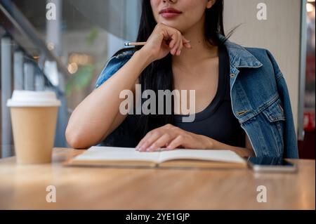 Eine Nahaufnahme einer schönen, nachdenklichen asiatischen Frau, die mit ihrem Tagebuch am Fenster in einem Café sitzt und etwas in Betracht zieht, während sie draußen blickt. Stockfoto
