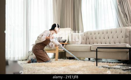 Eine glückliche, schöne asiatische Frau genießt Musik auf ihren Kopfhörern, während sie den Teppich im Wohnzimmer saugt. Reinigungsservice, Housekeeping, hous Stockfoto