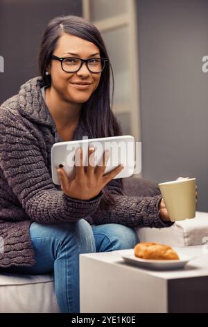 Junge Frau, die eine Brille mit ihrem digitalen Tablet trägt, während sie eine Tasse Kaffee trinkt und ein Croissant isst, bequem auf einer Couch sitzt Stockfoto