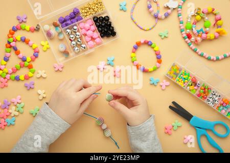 Das kleine Mädchen machte Armbänder auf beigefarbenem Hintergrund. Handgefertigter Schmuck mit Perlen für Kinder. Halsketten und Armbänder aus bunten Perlen und Perlen. DIY-BH Stockfoto