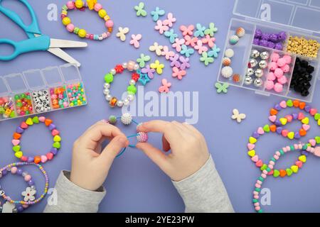 Das kleine Mädchen machte Armbänder auf lila Hintergrund. Handgefertigter Schmuck mit Perlen für Kinder. Halsketten und Armbänder aus bunten Perlen und Perlen. DIY br Stockfoto