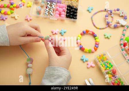 Das kleine Mädchen machte Armbänder auf beigefarbenem Hintergrund. Handgefertigter Schmuck mit Perlen für Kinder. Halsketten und Armbänder aus bunten Perlen und Perlen. DIY-BH Stockfoto