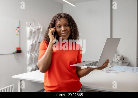 afroamerikanische, junge Schneiderin mit Laptop, die am Telefon über etwas spricht Stockfoto