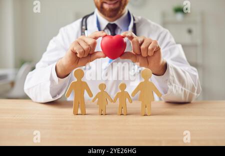 Mann Doktor, der das rote Herz in den Händen hält und Holzfiguren der Familie in der medizinischen Klinik schützt Stockfoto