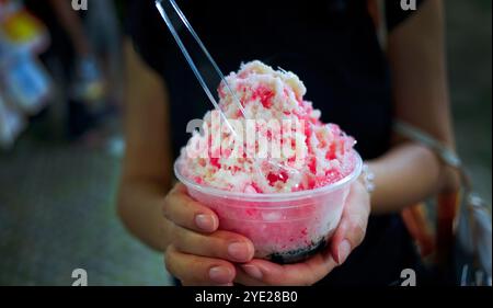 Weibliche Hände halten eine Plastikschale mit rotem Sirup rasiertes Eisdressing mit gesüßter Kondensmilchsoße darüber, beliebtes Dessert im Fun Fair Local Str Stockfoto