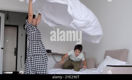 Eine Mutter schüttelt spielerisch eine Decke, während ihr Sohn eifrig aufs Bett springt. Diese fröhliche Szene fängt Familienverbundenheit und die einfachen Freuden von ein Stockfoto