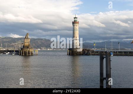 Leuchtturm Lindau und Bayerische Löwenstatue am Bodensee, mit Bergen, die in der Ferne unter bewölktem Himmel sichtbar sind Stockfoto