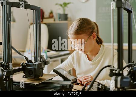 Ingenieurin arbeitet intensiv an einem Laptop, während sie im Büro von 3D-Druckern umgeben ist und ein neues Projekt erstellt. Fokus auf Technologie und Innovation in modernen Arbeitsumgebungen Stockfoto