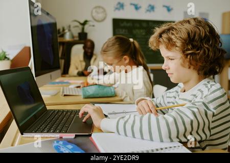 Gruppe von Kindern, die am Tisch lernen, mit Laptops und Notebooks, im Klassenzimmer, Schüler zeigen Fokus und Zusammenarbeit, vielfältige Umgebung Stockfoto