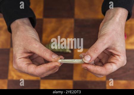 Nahaufnahme von Händen, die ein gemahlenes, mit Cannabis gefülltes Rollpapier verdrehen, um den Rollprozess der Herstellung eines Joint abzuschließen. Unkrautknospen auf dem Tisch unten Stockfoto