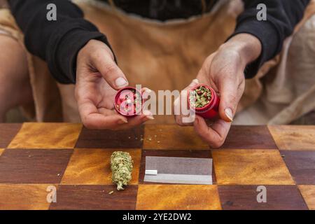 Selektive Nahaufnahme der Hände einer Person, die eine rote Unkraut gefüllte Mahlmaschine von oben hält. Karierter Tisch mit Papierrollen, Filter und Bud Disla Stockfoto