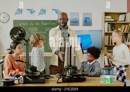 Afroamerikaner Lehrer unterrichtet verschiedene Gruppen von Schülern über 3D-Drucktechnologie im Unterricht. Kinder beobachten aufmerksam und engagieren sich am praktischen Lernen Stockfoto