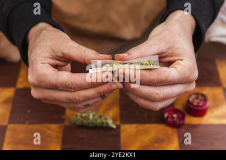 Nahaufnahme von Händen, die ein gemahlenes, mit Cannabis gefülltes Papierrollenpapier kneifen, um es in einen Joint zu Rollen. Unkrautknospen und Mahlwerk auf dem Tisch unten Stockfoto