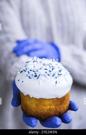 Die Hände des Kochs in blauen Handschuhen halten den Osterkuchen mit weißem Belag und blauen Streuseln. Frau mit traditionellem russischen Osterkuchen. Hausgemachter Kuchen Stockfoto