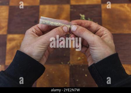 Ein großer Nahaufnahme von Fingern, die das Ende eines mit Cannabis gefüllten Papierrollenpapiers einklemmen und es zu einem Joint über einem karierten Tisch im Rücken Formen Stockfoto