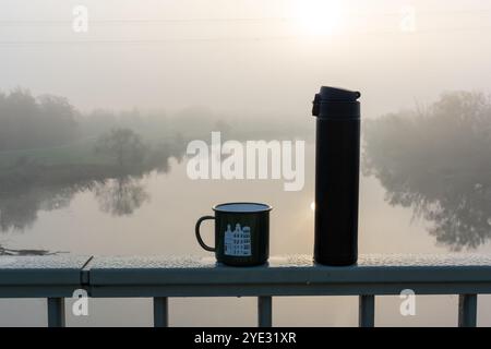 Thermoskanne und emaillierte Tasse auf frostigem Geländer mit Blick auf den nebeligen Fluss und die nebelige Landschaft bei Sonnenaufgang. Das Konzept der Ruhe, des Morgenrituals und der Wärme Stockfoto