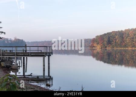 Ruhiger Herbstmorgen am Straussee, mit einem ruhigen Pier, der sich im stillen Wasser spiegelt, und Bäumen, die leuchtende Herbstfarben entlang der Küste zeigen Stockfoto