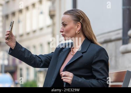 Eine wunderschöne 40-45-jährige Frau, die Selfies macht, sitzt auf einer Bank in der Stadt und trägt an einem sonnigen Tag einen stylischen Blazer mit schickem Haar. Stockfoto