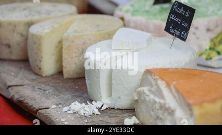 Eine Auswahl handwerklicher Käsesorten wird auf einem Holzbrett auf einem geschäftigen Markt in Alba, Italien, präsentiert. Jeder Käse ist einzigartig geformt Stockfoto