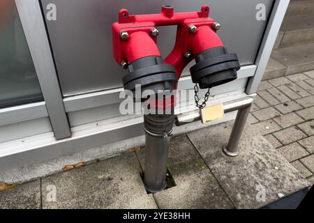 Ein hellroter Hydrantenanschluss befindet sich auf dem Bürgersteig in der Nähe eines Gebäudes und weist zwei separate Auslässe und eine gesicherte Kette auf, die an einer der Kappen befestigt ist. Stockfoto