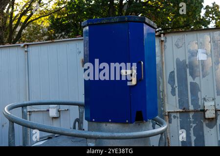 A blue control box secured with a lock is positioned on top of a utility pole. Surrounding trees and a gray fencing create a serene outdoor environment. Stockfoto
