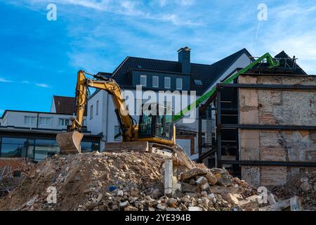 Ein Bagger arbeitet auf einer Baustelle und entfernt Schutt aus einer abgerissenen Struktur, während sich ein grüner Kran in der Nähe unter blauem Himmel befindet. Stockfoto