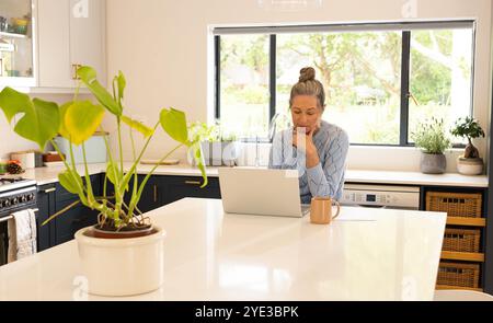 Reife Frau mit Laptop in heller Küche, konzentriert sich auf die Arbeit, zu Hause Stockfoto