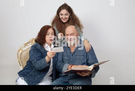 Ein Familienporträt von Eltern und einer erwachsenen Tochter, die ein Fotoalbum auf grauem Hintergrund betrachten. Stockfoto
