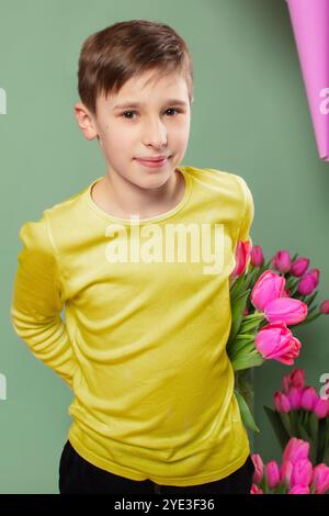 Der Junge versteckt Blumen hinter seinem Rücken und lächelt. Ein Kind mit einem Tulpenstrauß zum Muttertag. Stockfoto