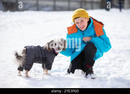 Ein Junge spielt draußen mit einem kleinen Hund im Winter. Posieren und auf die Kamera schauen. Ein Kind und ein Haustier auf einem Spaziergang. Stockfoto