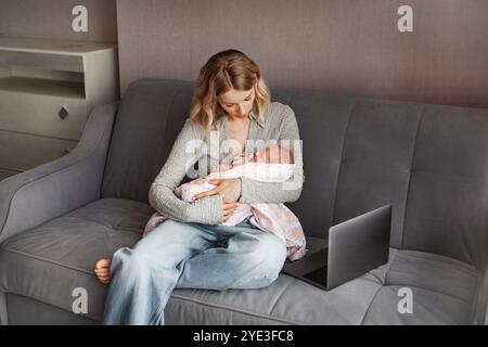 Arbeitende Mutter mit schlafendem Neugeborenen in den Armen, Multitasking mit dem Laptop, während sie zu Hause auf der Couch sitzt und Karriere und Mutterschaft miteinander in Einklang bringt Stockfoto