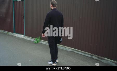 Ein Mann in einer schwarzen Jacke und Mütze läuft eine ruhige Straße entlang, ein burgunderroter Metallzaun im Hintergrund. Street Style wird mit dem Konzept von kombiniert Stockfoto