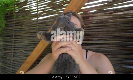 Eine junge Frau hält ein Tier in den Armen. Ein ruhiges graues Kaninchen, das von einer Person auf einem Holztisch im Freien gepflegt wird. Stockfoto