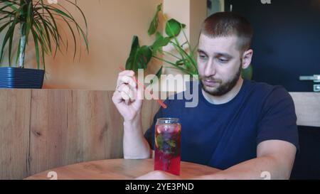 Blumen in Vasen im Hintergrund schaffen eine ruhige Atmosphäre für diesen erfrischenden Moment. Der junge Mann spürt den Sommer, wenn er einen kühlen Cocktail schlürft Stockfoto