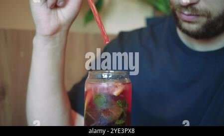 Blumen in Vasen im Hintergrund schaffen eine ruhige Atmosphäre für diesen erfrischenden Moment. Der junge Mann spürt den Sommer, wenn er einen kühlen Cocktail schlürft Stockfoto