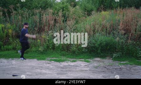 Ein Mann im schwarzen Trainingsanzug erkundet einen Naturwanderweg, der von wilden Pflanzen und Bäumen umgeben ist. Geeignet für Natur-, Abenteuer- und Outdoor-Videos. Stockfoto