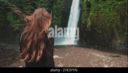 Eine junge Frau mit langen braunen Haaren genießt einen wunderschönen Wasserfall in einem tropischen Dschungel-Wald. Der Wasserfall stürzt über eine felsige Klippe, umgeben von üppiger grüner Vegetation Stockfoto