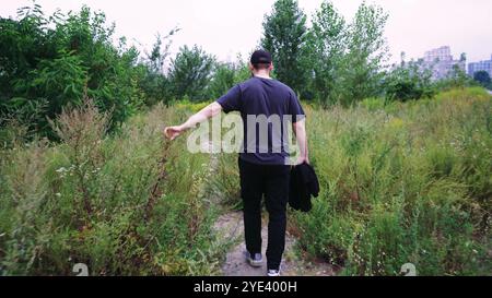 Ein Mann im schwarzen Trainingsanzug erkundet einen Naturwanderweg, der von wilden Pflanzen und Bäumen umgeben ist. Geeignet für Natur-, Abenteuer- und Outdoor-Videos. Stockfoto