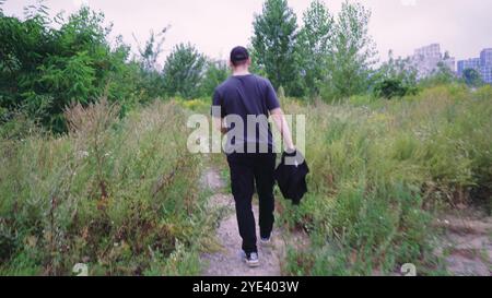 Ein Mann im schwarzen Trainingsanzug erkundet einen Naturwanderweg, der von wilden Pflanzen und Bäumen umgeben ist. Geeignet für Natur-, Abenteuer- und Outdoor-Videos. Stockfoto