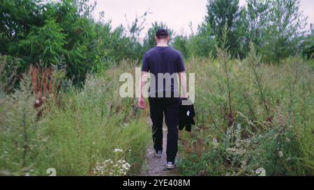 Ein Mann im schwarzen Trainingsanzug erkundet einen Naturwanderweg, der von wilden Pflanzen und Bäumen umgeben ist. Geeignet für Natur-, Abenteuer- und Outdoor-Videos. Stockfoto