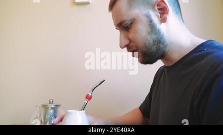 Ein Wasserkocher dampft auf dem Tisch, die Atmosphäre ist ruhig und entspannt. Ein Mann trinkt Tee aus einem traditionellen Keramikbecher, tief in Gedanken. Stockfoto