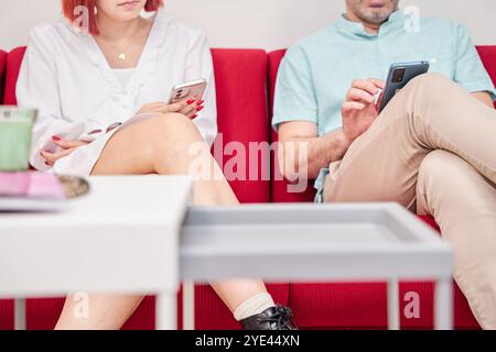 Unerkennbares Paar, das auf dem Sofa sitzt, in einem Wartezimmer mit seinem Smartphone Stockfoto