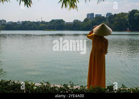 Hanoi, Vietnam: Vietnamesische Frau, die vor der Sonne mit ihrem traditionellen konischen Hut von Non La schützt, vor dem Hoan Kiem See (Schwertsee), Altstadt Stockfoto