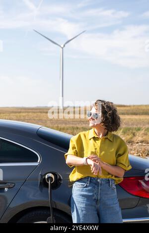 Eine Frau mit gewölbten Haaren, die ein gelbes Hemd und eine Sonnenbrille trägt, steht neben dem Aufladen des Elektroautos. Windräder im Hintergrund. Stockfoto