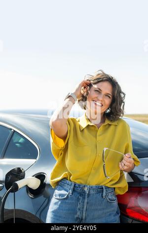 Frau mit gebogenen Haaren, die ein gelbes Hemd trägt, steht neben dem Aufladen des Elektroautos. Windräder im Hintergrund. Stockfoto