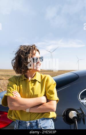 Eine Frau mit gewölbten Haaren, die ein gelbes Hemd und eine Sonnenbrille trägt, steht neben dem Aufladen des Elektroautos. Windräder im Hintergrund. Stockfoto