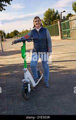 Eine Frau, die neben einem Elektroroller auf einem gepflasterten Weg steht. Sie trägt einen grauen Pullover und eine marineblaue Weste und lächelt in die Kamera. Der Hintergrund Stockfoto