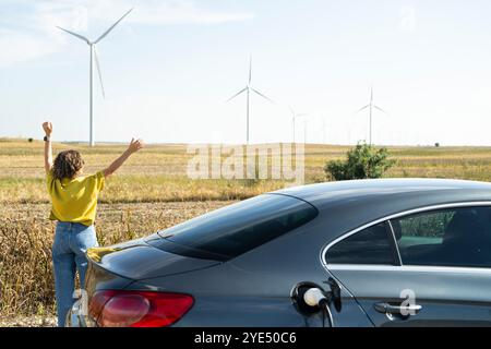 Frau mit gebogenen Haaren, die ein gelbes Hemd trägt, steht neben dem Aufladen des Elektroautos. Windräder im Hintergrund. Stockfoto