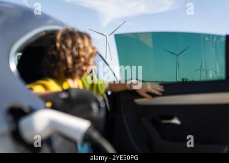 Eine Frau mit gebogenen Haaren, die ein gelbes Hemd trägt, sitzt in einem Elektroauto. Windräder im Hintergrund. Stockfoto