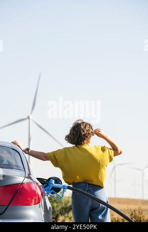 Die Frau hält eine Wasserstoff-Tankdüse. Betanken des Autos mit Wasserstoffkraftstoff. Windräder im Hintergrund. Stockfoto